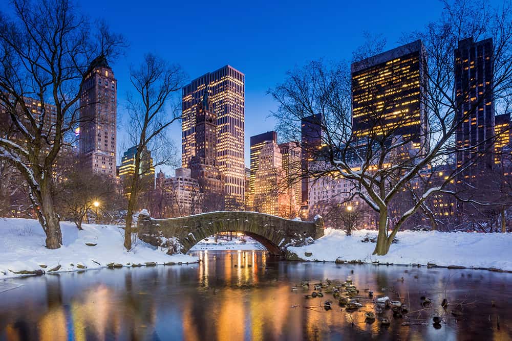 Công viên Central Park phủ đầy tuyết trắng lúc hoàng hôn nổi bật với cây cầu đá bắc qua một hồ nước tĩnh lặng, bao quanh là những hàng cây trơ trụi. Những tòa nhà chọc trời được thắp sáng rực rỡ chiếu rọi bầu trời mùa đông.