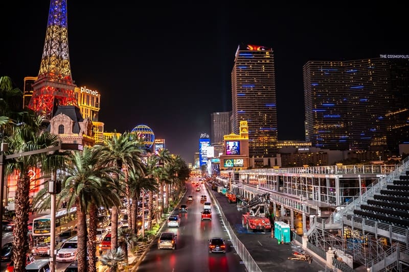 Dải Las Vegas nhộn nhịp về đêm, với những biển hiệu rực rỡ ánh đèn, hàng cây cọ và giao thông tấp nập. Ánh đèn màu sắc tạo nên một bầu không khí sống động và tràn đầy năng lượng.