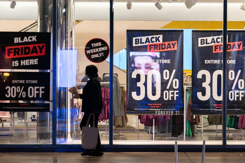Một người đi ngang qua cửa sổ một cửa hàng có biển hiệu lớn "Black Friday" quảng cáo giảm giá 30% cho toàn bộ cửa hàng. Không khí nhộn nhịp và đậm chất quảng cáo.