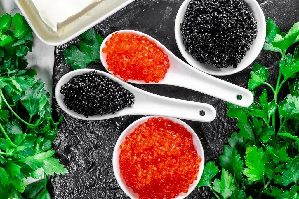 Trứng cá muối các loại trên thìa trắng: những viên ngọc trai đỏ và đen rực rỡ nổi bật trên nền tối màu, được trang trí bằng rau mùi tây tươi và một khối bơ.