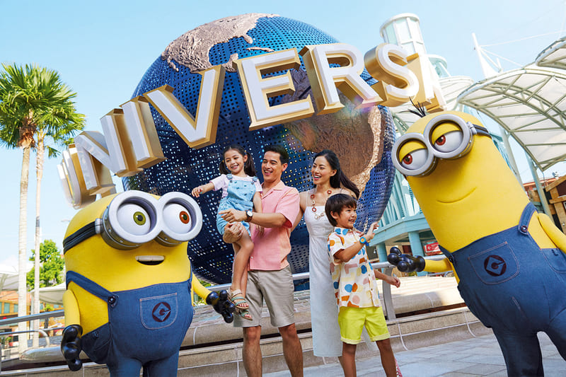 Một gia đình tươi cười rạng rỡ bên hai nhân vật "Minions" tại lối vào công viên giải trí. Phía sau là quả địa cầu với dòng chữ "UNIVERSAL" cỡ lớn. Một ngày nắng đẹp.