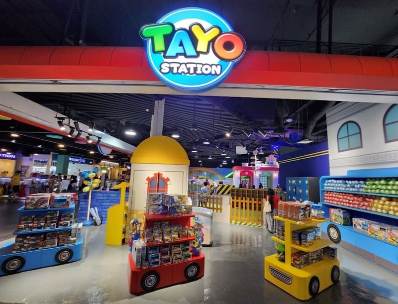 Khu vui chơi trong nhà đầy màu sắc với biển hiệu "Tayo Station" phía trên lối vào. Xe đồ chơi, hình ảnh rực rỡ và các hoạt động dành cho trẻ em tạo nên một bầu không khí sống động và vui vẻ.