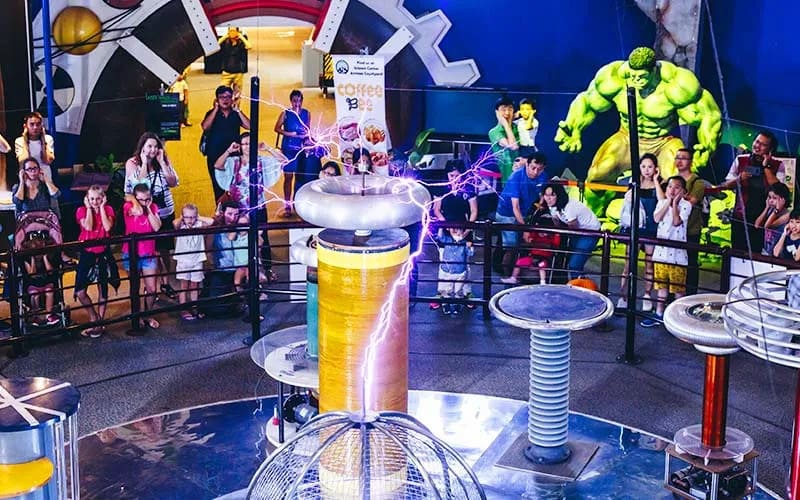 Một nhóm người, trong đó có cả trẻ em, đang quan sát cuộn dây Tesla phát ra tia lửa màu tím tại một bảo tàng khoa học. Một bức tượng siêu anh hùng màu xanh lá cây khổng lồ đứng ở phía sau.