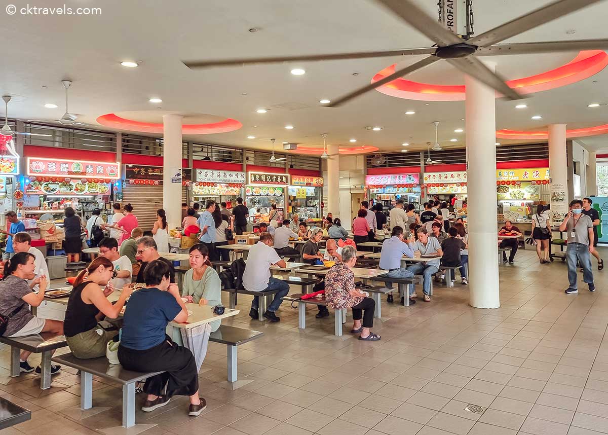 Một khu ẩm thực nhộn nhịp với nhiều quầy hàng bán đồ ăn, biển hiệu rực rỡ và nhiều người ngồi ở bàn thưởng thức bữa ăn. Không khí sống động và thân thiện.