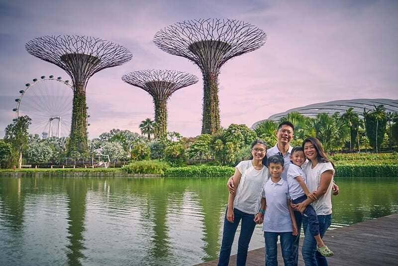 Một gia đình năm người, tươi cười, đứng bên một dòng kênh yên tĩnh tại Gardens by the Bay, Singapore. Những cây cổ thụ khổng lồ và vòng đu quay hiện lên nổi bật trong khung cảnh xanh tươi, thơ mộng phía sau.