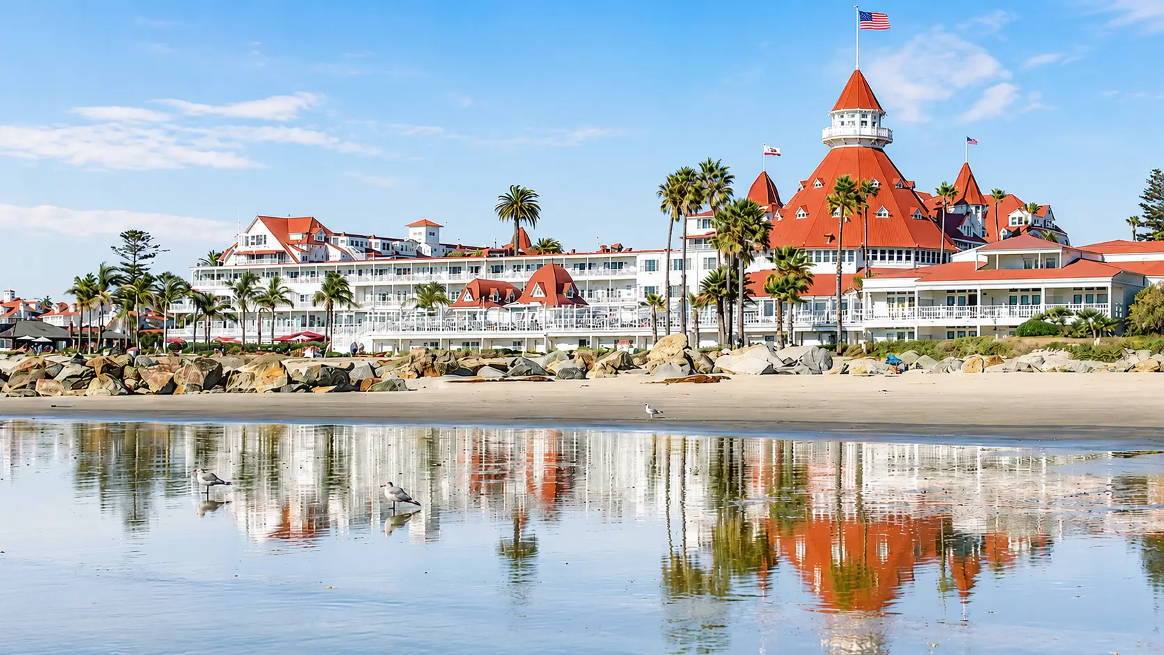 Khách sạn Hotel del Coronado với kiến trúc trắng, mái ngói đỏ đặc trưng bên bãi biển California, phản chiếu trên mặt nước dưới bầu trời xanh trong.