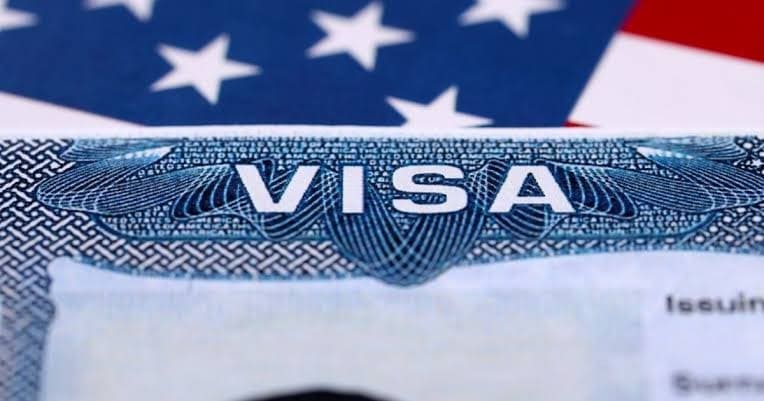 Tờ visa Mỹ cận cảnh với chữ VISA in nổi bật trên nền hoa văn màu xanh, phía sau là quốc kỳ Mỹ