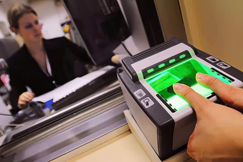 Ngón tay đặt trên máy quét vân tay phát ánh sáng xanh tại quầy làm thủ tục có nhân viên ngồi phía sau