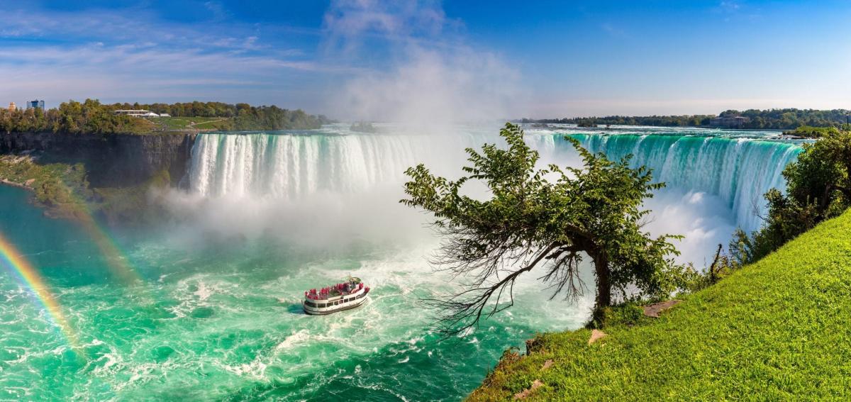 Một khung cảnh sống động của thác Niagara với một chiếc thuyền du lịch đang lướt trên mặt nước màu ngọc lam mờ ảo. Cầu vồng uốn cong ở bên trái, và thảm thực vật xanh tươi trải dài dọc theo bờ bên phải.