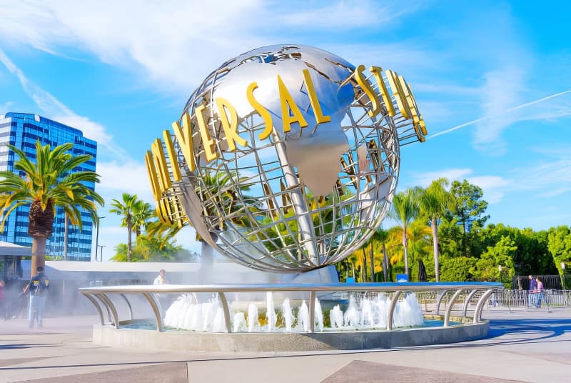 Một quả cầu kim loại lớn có logo "Universal Studios" được đặt ở trung tâm giữa những cây cọ và đài phun nước, dưới bầu trời xanh trong vắt, tạo nên một khung cảnh sống động.