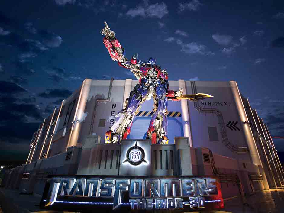 Optimus Prime, một người máy khổng lồ, đứng oai vệ trước một tòa nhà lúc hoàng hôn, chỉ tay lên trời. Biển hiệu ghi "Transformers The Ride 3D".