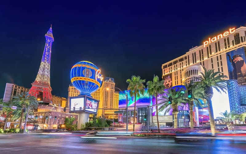Dải Las Vegas rực rỡ về đêm, với mô hình tháp Eiffel và những ánh đèn neon đầy màu sắc. Không khí sôi động với những hàng cây cọ và đường phố nhộn nhịp.