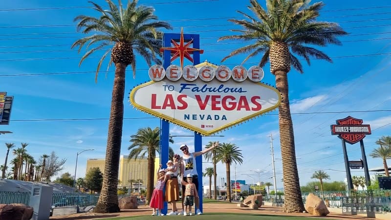 Một gia đình vui vẻ tạo dáng trước tấm biển nổi tiếng "Chào mừng đến với Las Vegas tuyệt vời, Nevada". Những hàng cây cọ tô điểm thêm cho khung cảnh dưới bầu trời xanh trong vắt.