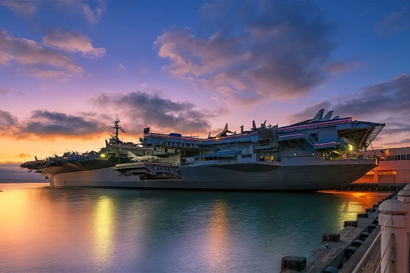 Tàu sân bay neo đậu lúc hoàng hôn dưới bầu trời rực rỡ. Mặt nước phản chiếu những sắc cam và tím, tạo nên một khung cảnh thanh bình nhưng hùng vĩ tại bến cảng.