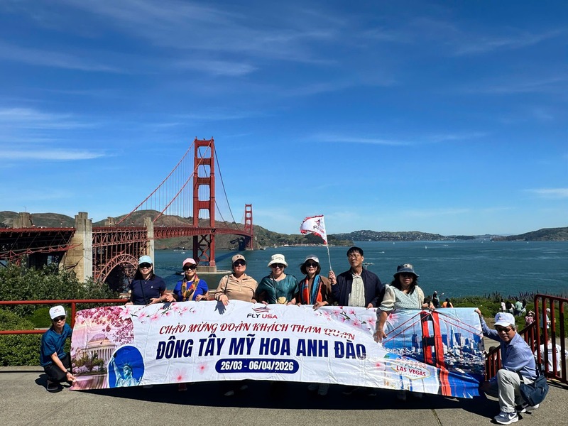 Một nhóm người đứng trước cầu Cổng Vàng, cầm một biểu ngữ. Biểu ngữ có ghi thông tin du lịch bằng tiếng Việt. Bầu trời trong xanh.