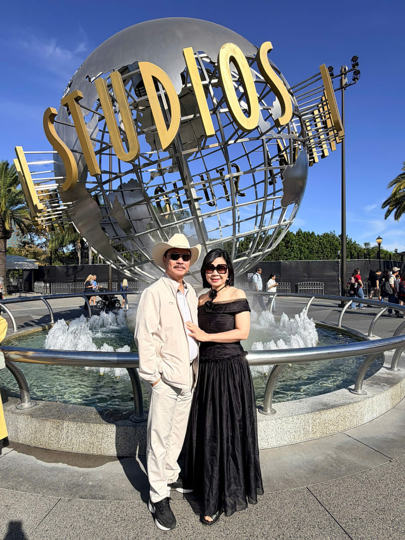 Một cặp đôi đứng mỉm cười trước quả cầu biểu tượng của Universal Studios. Người đàn ông đội mũ cao bồi và mặc trang phục màu be; người phụ nữ mặc váy đen. Trời nắng đẹp.
