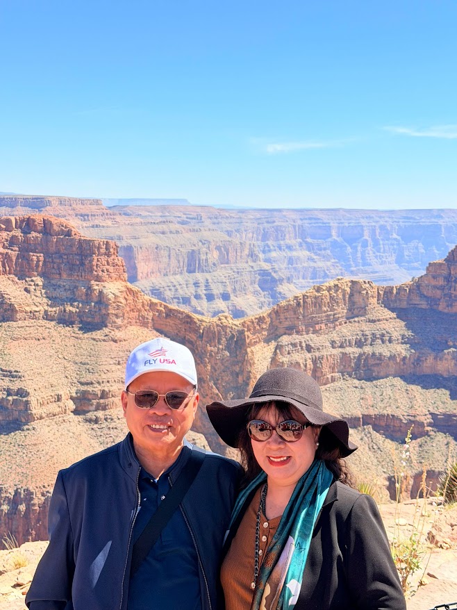 Một cặp đôi tươi cười đứng trước hẻm núi Grand Canyon rộng lớn, ngập tràn ánh nắng. Cả hai đều đeo kính râm và đội mũ; bầu trời trong xanh, toát lên cảm giác vui vẻ và phiêu lưu.