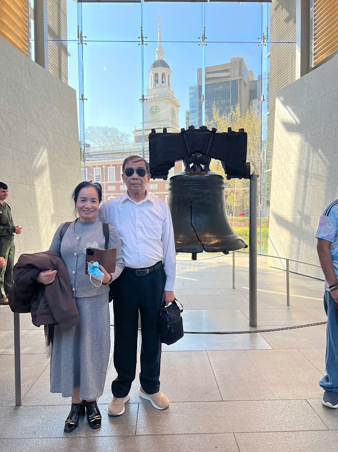 Một cặp đôi tươi cười đứng trước Chuông Tự do bên trong một bảo tàng ngập tràn ánh nắng, với Tòa nhà Độc lập hiện ra qua những cửa sổ kính lớn ở phía sau.