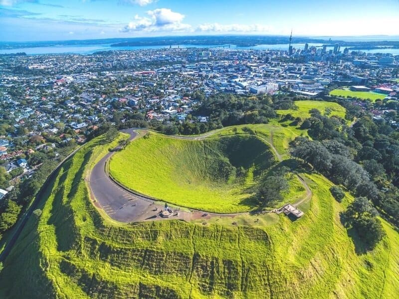 Toàn cảnh từ trên cao miệng núi lửa Mount Eden phủ cỏ xanh và thành phố Auckland với tháp Sky Tower phía xa