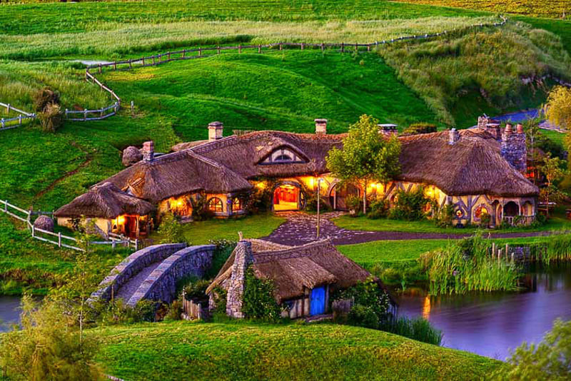 Quán trọ Green Dragon mái tranh sáng đèn bên cầu đá và hồ nước giữa đồi cỏ xanh ở làng Hobbiton