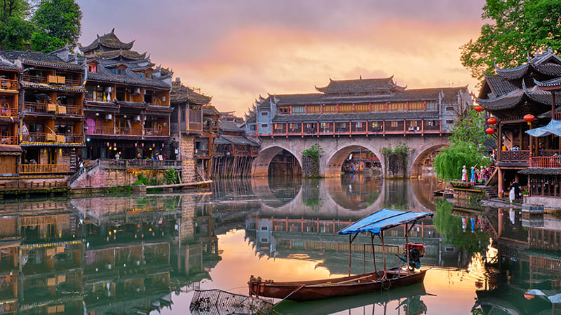 Phượng Hoàng cổ trấn với thuyền gỗ trên sông Đà Giang, phía xa là cầu đá vòm và dãy nhà sàn gỗ phản chiếu xuống mặt nước