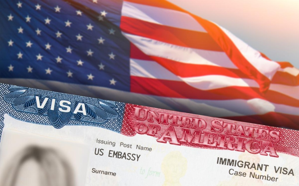Một phần thị thực Hoa Kỳ hiện ra ở phía trước với dòng chữ "United States of America" (Hợp chủng quốc Hoa Kỳ). Lá cờ Mỹ đang tung bay ở phía sau.