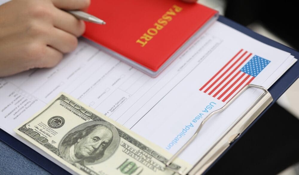 Một cuốn hộ chiếu màu đỏ được đặt phía trên một mẫu đơn xin visa có hình cờ Mỹ. Một tờ tiền trăm đô la nằm trên một tấm bảng kẹp giấy, cho thấy sự chuẩn bị cho chuyến đi.