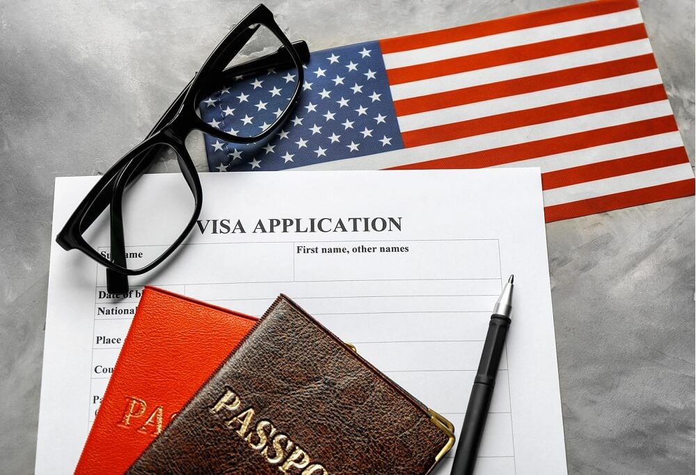 Tờ đơn xin visa cùng hai hộ chiếu, kính mắt, bút và quốc kỳ Mỹ trên mặt bàn