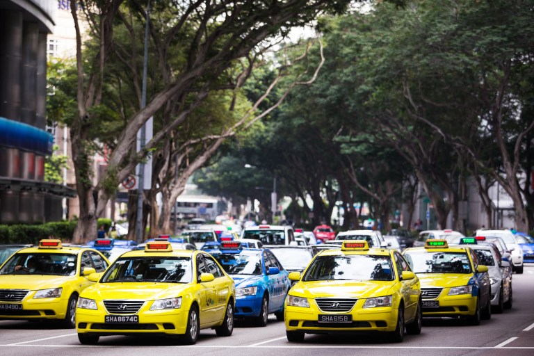 Một con phố đô thị nhộn nhịp với những hàng cây cao rợp bóng, những chiếc taxi màu vàng và xanh lam chạy san sát nhau trên nhiều làn đường. Khung cảnh này thể hiện sự chuyển động và nhịp sống thành thị.