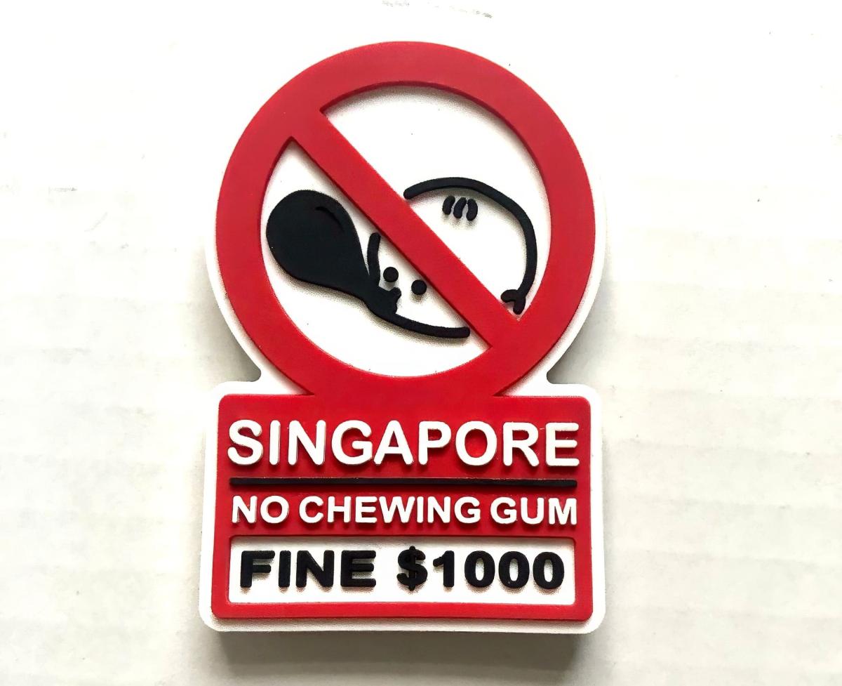 Biển báo màu đỏ trắng có hình biểu tượng kẹo cao su bị cấm kèm dòng chữ: "Singapore Cấm nhai kẹo cao su, phạt 1000 đô la Singapore." Thể hiện quy định nghiêm ngặt.