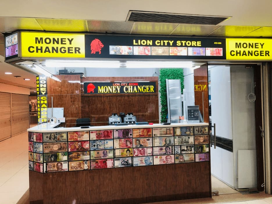 Một quầy đổi tiền mang tên "Lion City Store" trưng bày nhiều loại tiền giấy quốc tế. Biển hiệu và ánh đèn sáng tạo nên không gian chuyên nghiệp.
