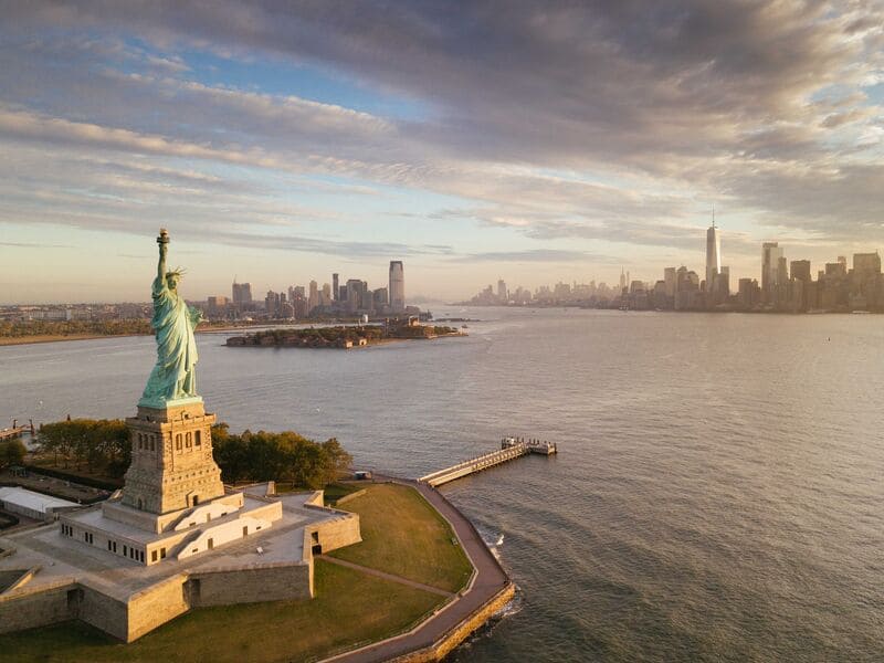 Ảnh chụp từ trên cao tượng Nữ thần Tự do với đường chân trời thành phố New York ở phía xa lúc hoàng hôn. Bầu trời nhiều mây, tạo nên một sắc vàng rực rỡ cho khung cảnh.