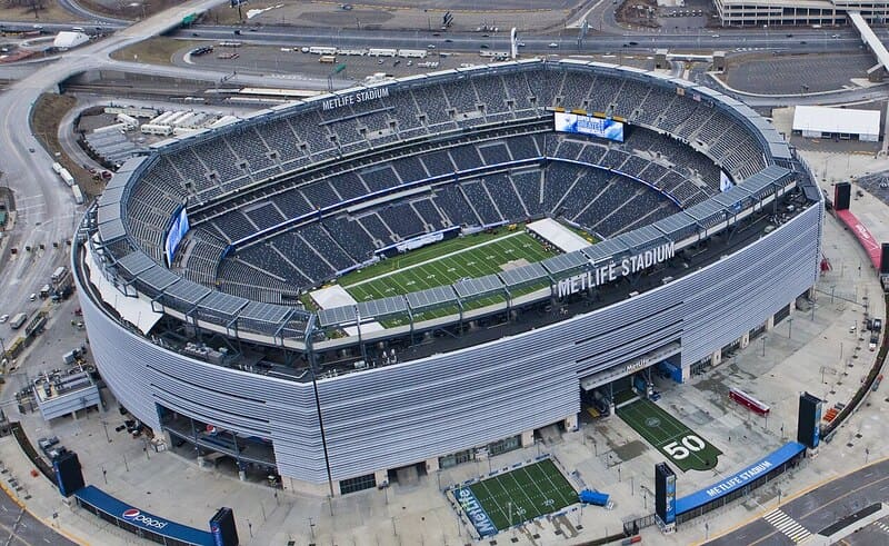 Ảnh chụp từ trên cao sân vận động MetLife, một đấu trường hình bầu dục rộng lớn, trống trải với thảm cỏ xanh mướt, bao quanh là những hàng ghế màu xám và đường phố đô thị, dưới bầu trời nhiều mây.