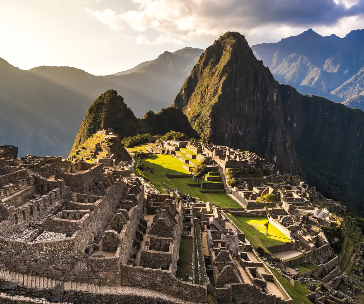 Hình ảnh từ trên cao của Machu Picchu lúc bình minh, với những công trình đá cổ kính, những thửa ruộng bậc thang xanh mướt và những ngọn núi hùng vĩ, tạo nên một bầu không khí thanh bình và đậm chất lịch sử.