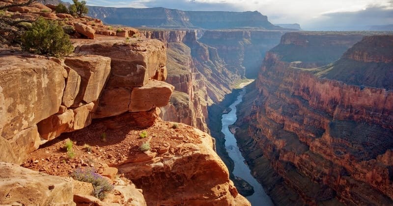 Quang cảnh Grand Canyon với những vách đá đỏ gồ ghề và cây cối thưa thớt. Sông Colorado uốn lượn qua thung lũng sâu dưới bầu trời nhiều mây, gợi lên vẻ đẹp hùng vĩ.
