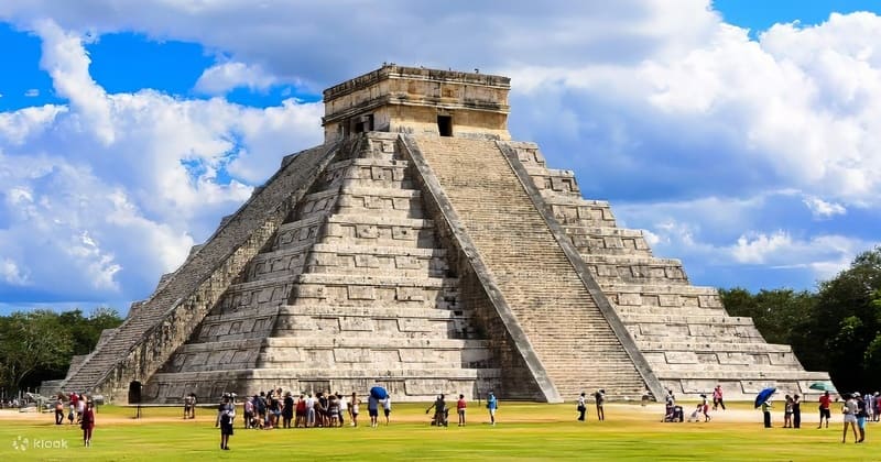 Một kim tự tháp đá lớn, El Castillo ở Chichén Itzá, dưới bầu trời xanh trong vắt với những đám mây trắng bồng bềnh. Du khách tập trung dưới chân kim tự tháp, ghi lại vẻ đẹp lịch sử hùng vĩ của nó.