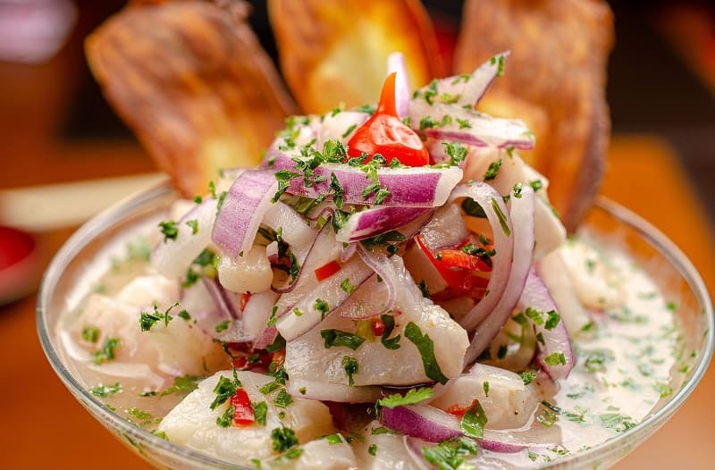Hình ảnh cận cảnh món ceviche tươi ngon trong bát thủy tinh, với cá thái hạt lựu, hành tây đỏ, rau mùi và ớt đỏ. Chuối chiên trang trí phía sau, tạo nên một vẻ ngoài đầy màu sắc và tươi mới.