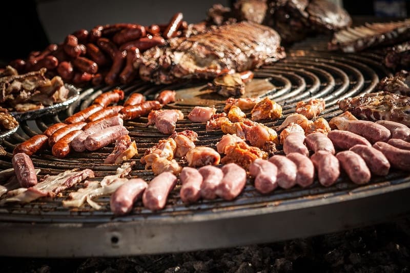 Nhiều loại thịt, bao gồm xúc xích, sườn và thịt gà, đang xèo xèo trên một vỉ nướng tròn lớn. Khung cảnh toát lên không khí sôi động của một buổi tiệc nướng.