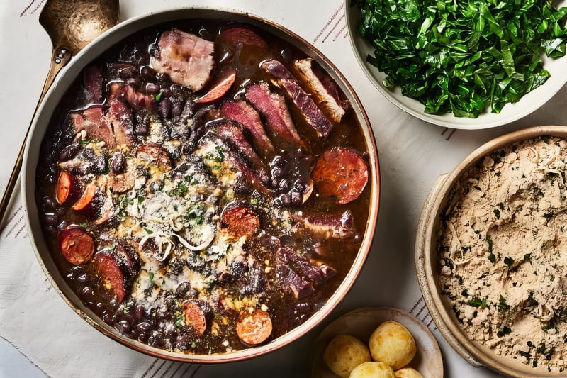 Một món feijoada Brazil đầy màu sắc với đậu đen, xúc xích và các loại thịt. Đi kèm là các bát rau xanh thái nhỏ, farofa và bánh mì nhỏ.
