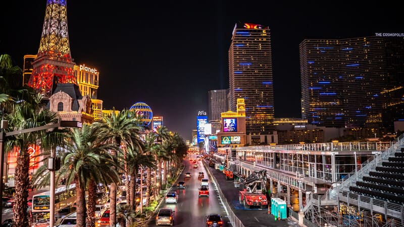 Cảnh đêm của dải Las Vegas Strip, nhộn nhịp xe cộ và được chiếu sáng bởi những ánh đèn neon rực rỡ. Những hàng cây cọ trải dọc đường phố, càng làm tăng thêm bầu không khí sống động.