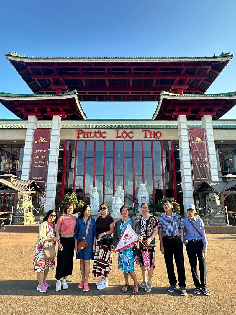 Một nhóm tám người đứng mỉm cười trước một tòa nhà lớn có điểm nhấn màu đỏ và các bức tượng, dưới tấm biển ghi "Phúc Lộc Thọ". Bầu trời trong xanh.