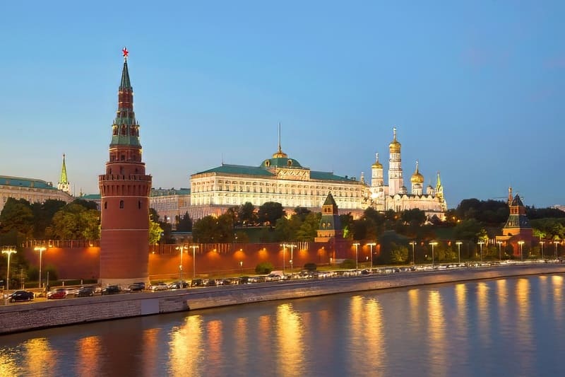 Khung cảnh Điện Kremlin lúc hoàng hôn với những mái vòm vàng óng, tường gạch đỏ và hình ảnh phản chiếu của dòng sông dưới bầu trời trong xanh, gợi lên cảm giác về sự tráng lệ lịch sử.