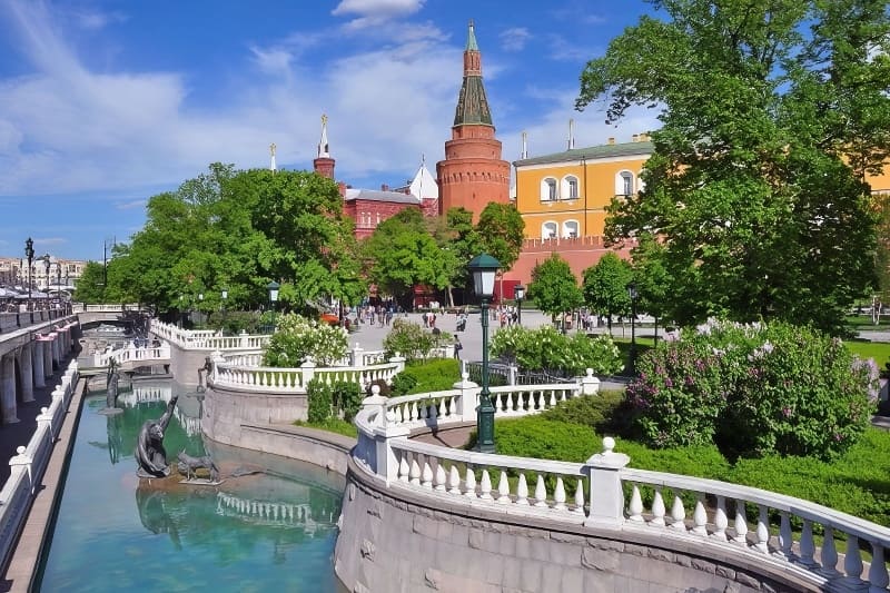 Khung cảnh công viên ngập nắng với kênh đào màu ngọc lam và các tác phẩm điêu khắc, bao quanh là cây xanh tươi tốt và những tòa nhà rực rỡ sắc màu. Mọi người tận hưởng một ngày trong lành, tươi sáng.