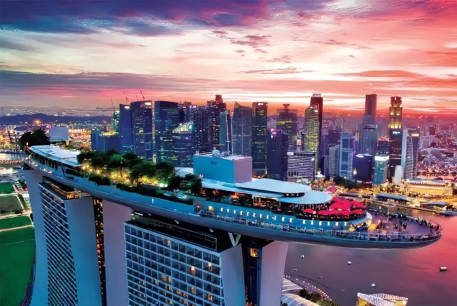 Hình ảnh từ trên cao của Marina Bay Sands lúc hoàng hôn, nổi bật với khu vườn và hồ bơi trên sân thượng mang tính biểu tượng. Toàn cảnh thành phố được chiếu sáng, tạo nên một khung cảnh sống động.