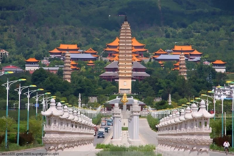 Một cây cầu lớn, được trang trí công phu dẫn đến một quần thể đền thờ rộng lớn với nhiều tháp và mái đỏ, nằm giữa khung cảnh núi non xanh tươi, gợi lên sự thanh bình.