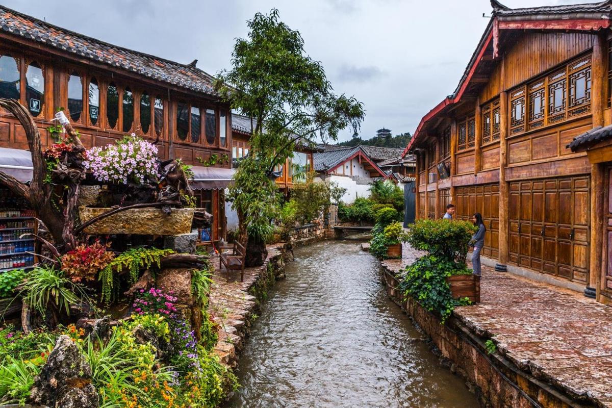 Một con kênh nhỏ chảy giữa những tòa nhà gỗ truyền thống với những chi tiết chạm khắc tinh xảo, được bao quanh bởi cây xanh tươi tốt và những bông hoa rực rỡ, tạo nên một bầu không khí thanh bình.