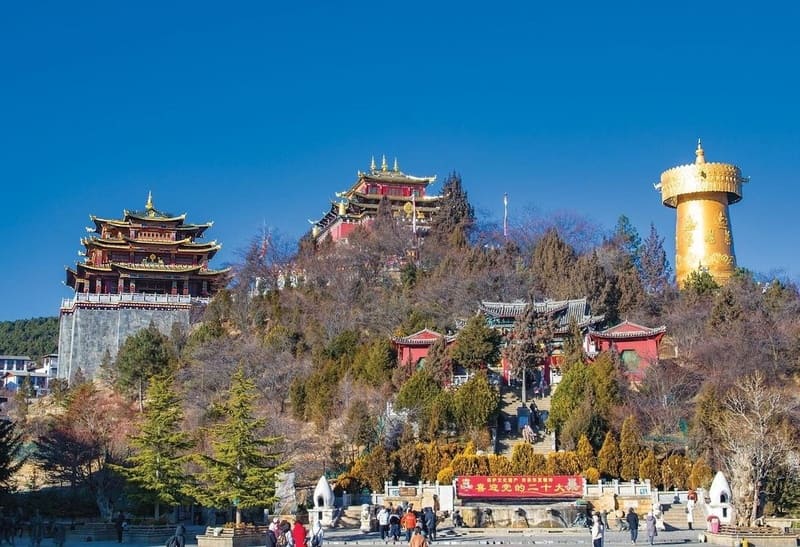 Một quần thể chùa Phật giáo nằm trên một ngọn đồi dưới bầu trời xanh trong vắt, mang kiến ​​trúc truyền thống Trung Quốc với những mái nhà trang trí công phu và một bánh xe cầu nguyện lớn bằng vàng.