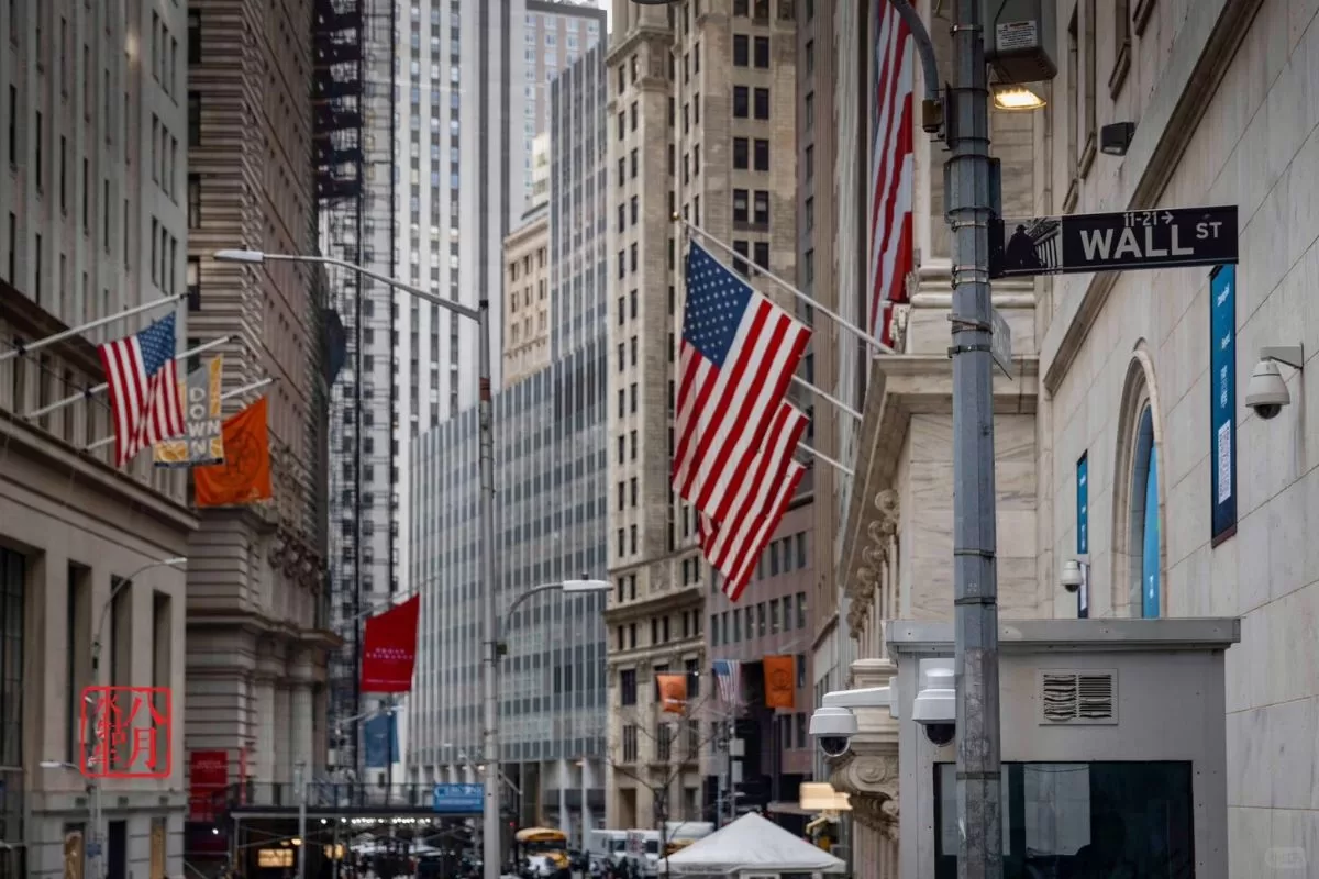 Cảnh đường phố phố Wall, New York, với những lá cờ Mỹ tung bay kiêu hãnh trên các tòa nhà. Những tòa nhà chọc trời cao vút nằm dọc theo con phố hẹp, tạo nên cảm giác về một bầu không khí tài chính nhộn nhịp.