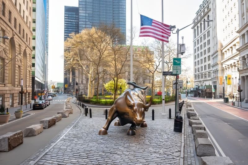 Tượng bò tót bằng đồng trên con phố lát đá cuội ở khu tài chính, thành phố New York. Lá cờ Mỹ tung bay phía trên, phía sau là những tòa nhà chọc trời và cây cối.