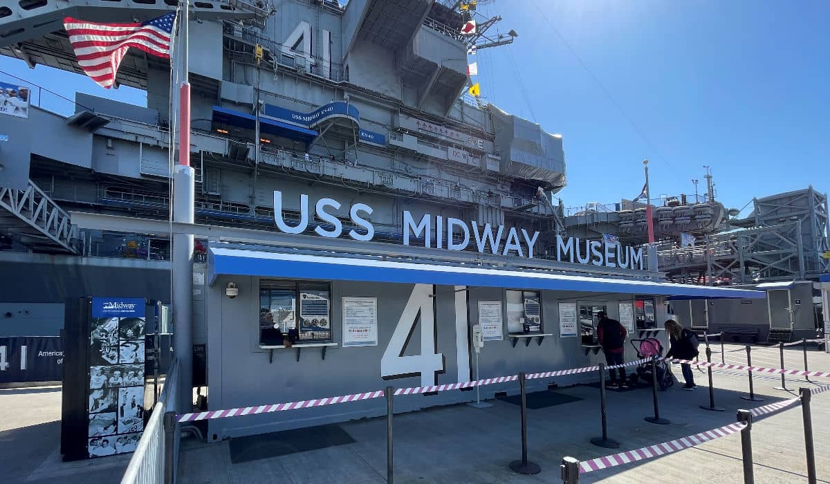 Lối vào Bảo tàng USS Midway với số "41" hiện rõ, mái che màu xanh lam và mọi người đang xếp hàng. Cờ bay phấp phới phía trên, tạo nên bầu không khí lịch sử, hải quân.